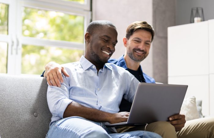 Gay,Couple,Using,Computer,For,Video,Conference,On,Sofa,Or